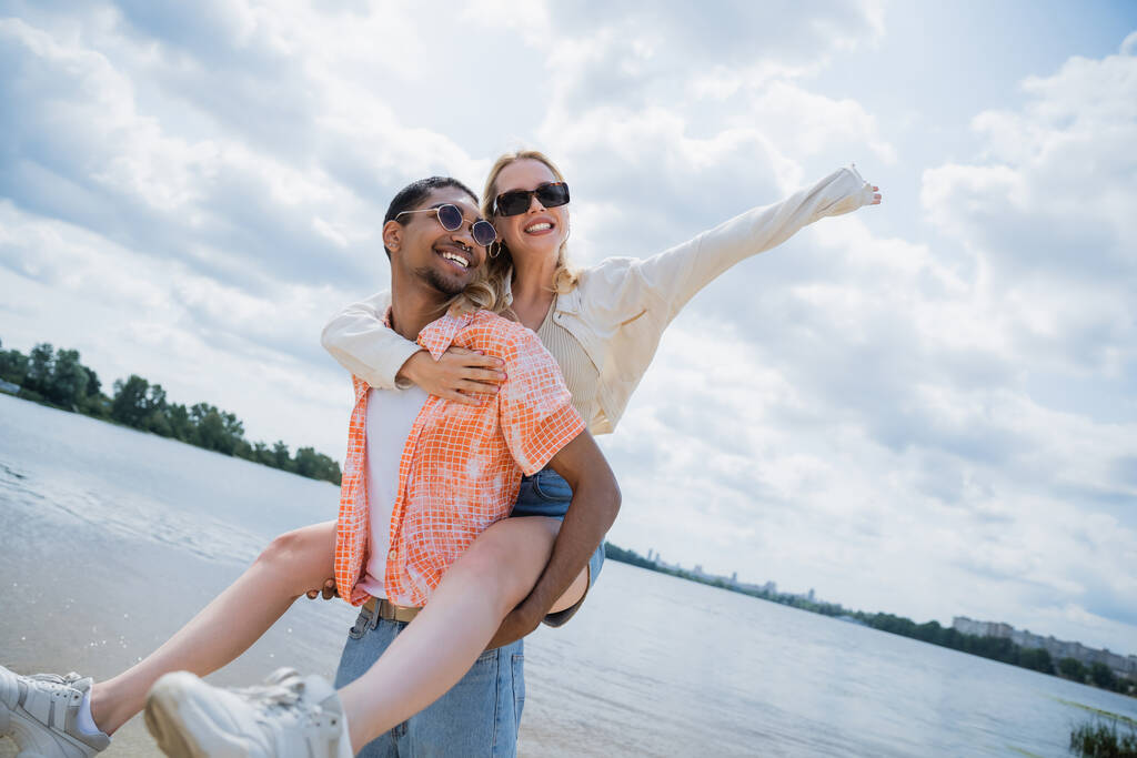 joyful african american man piggybacking happy woman with outstretched hand on riverside - Foto, immagini