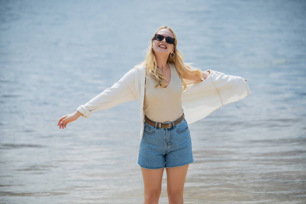 smiling blonde woman in denim shorts standing near river with outstretched hands - Photo, image