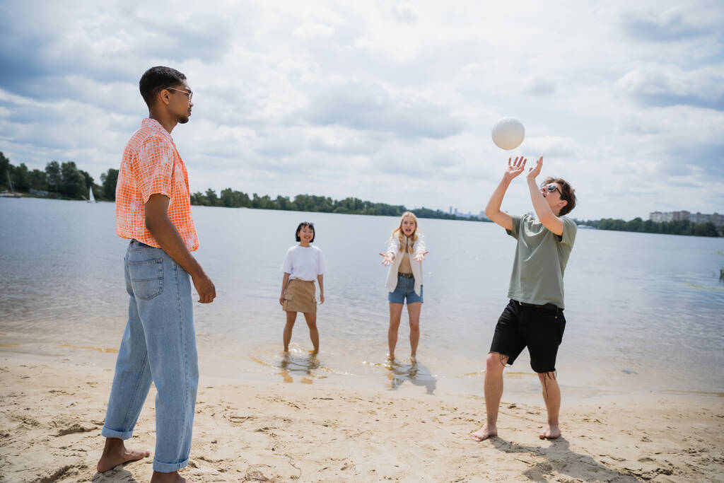 νεαρός άνδρας πασάρει μπάλα, ενώ παίζει βόλεϊ στην παραλία με διαφυλετικούς φίλους - Φωτογραφία, εικόνα
