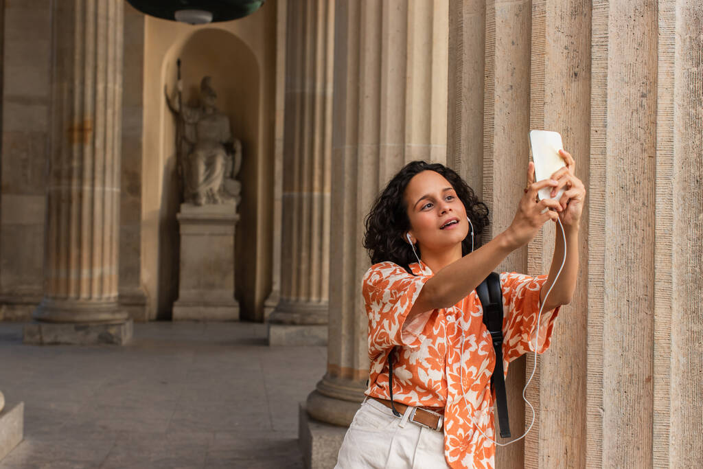 кудрявая молодая женщина в проводных наушниках делает селфи со статуей, держа в руках смартфон  - Фото, изображение