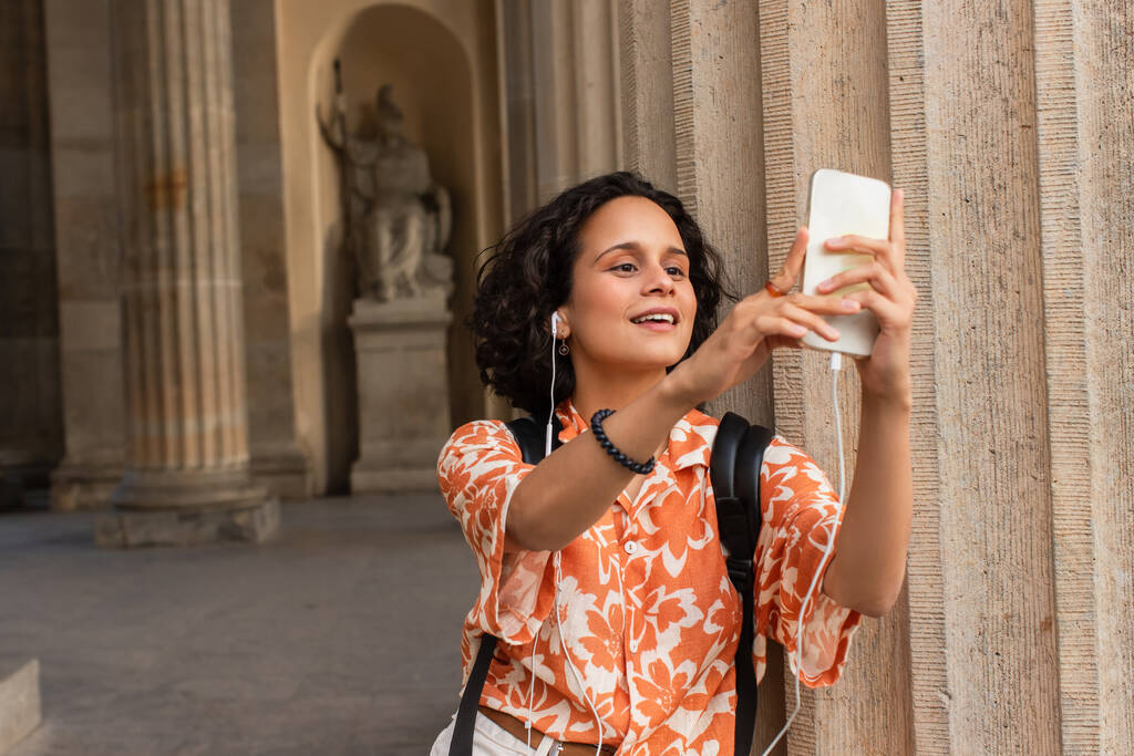 happy young tourist in wired earphones taking selfie with statue while holding smartphone  - Foto, Imagem