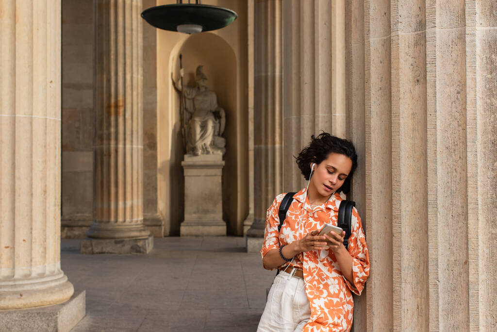 pretty young woman in wired earphones listening music and holding smartphone near statue in berlin  - Foto, Bild