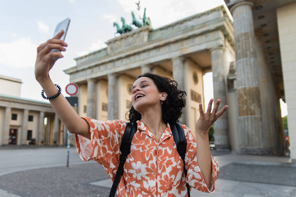 curly tourist with backpack taking selfie on smartphone near brandenburg gate in berlin  - Fotó, kép