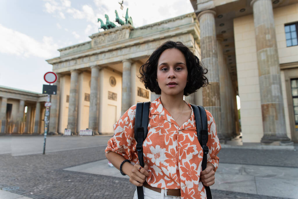 σγουρή νεαρή γυναίκα με σακίδιο στέκεται κοντά στην πύλη Brandenburg στο Βερολίνο  - Φωτογραφία, εικόνα