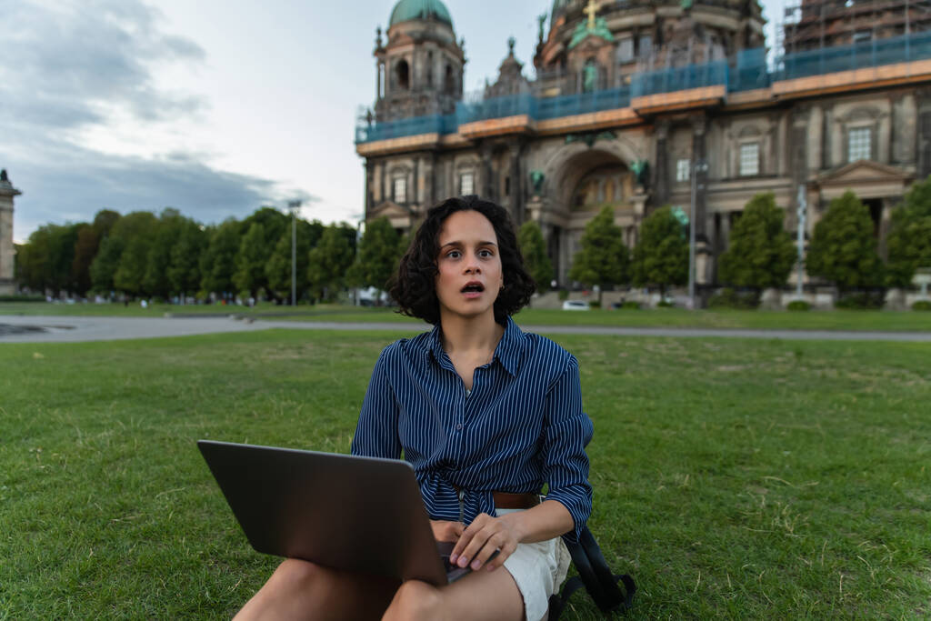 σοκαρισμένη νεαρή γυναίκα κρατώντας φορητό υπολογιστή και κάθεται στο γρασίδι κοντά σε θολή καθεδρικό ναό στο Βερολίνο  - Φωτογραφία, εικόνα