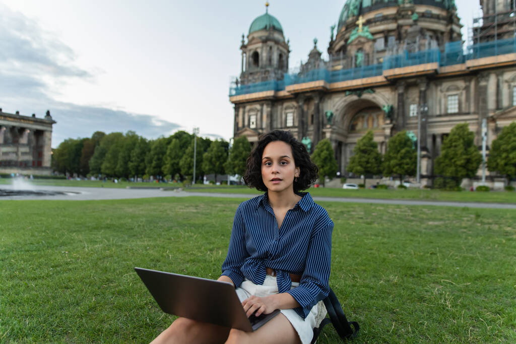 молодий фрілансер за допомогою ноутбука і сидячи на траві біля розмитого собору в Берліні під час подорожі.  - Фото, зображення