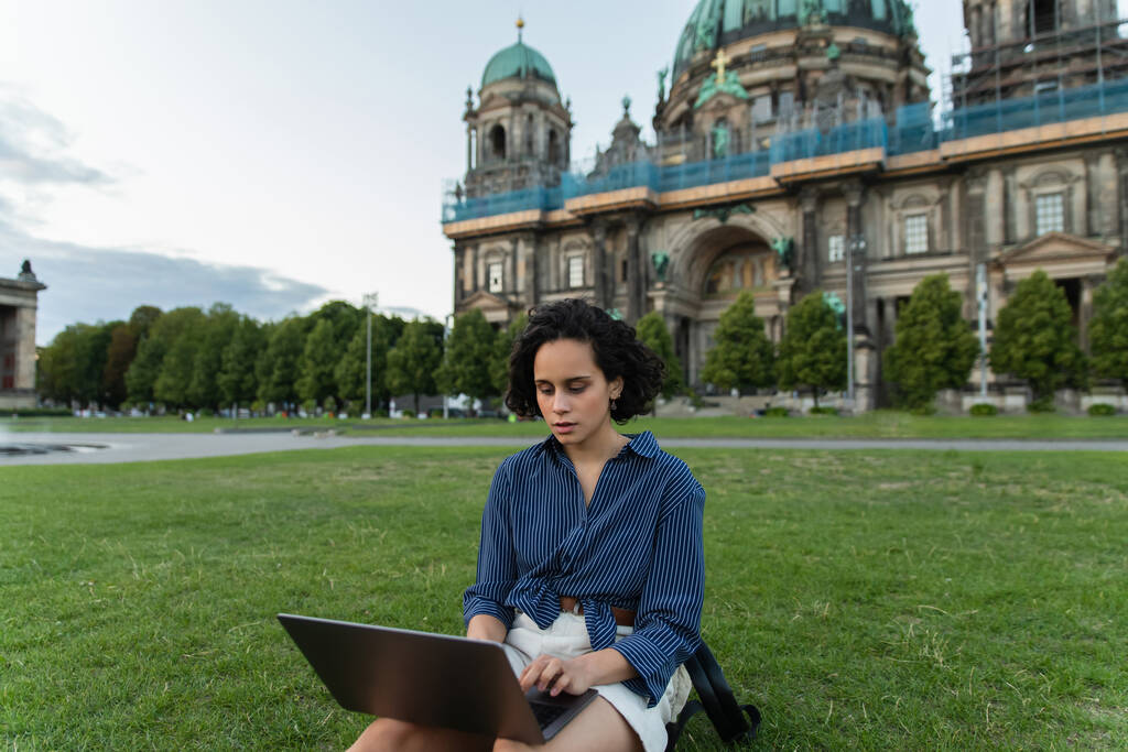 під час подорожі молода жінка використовує ноутбук і сидить на траві біля розмитого собору в берліні.  - Фото, зображення