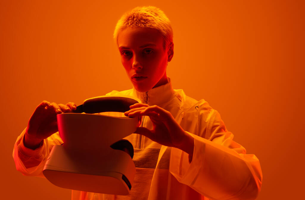 Young woman in futuristic raincoat putting on VR glasses and looking at camera under bright orange neon light - Fotó, kép