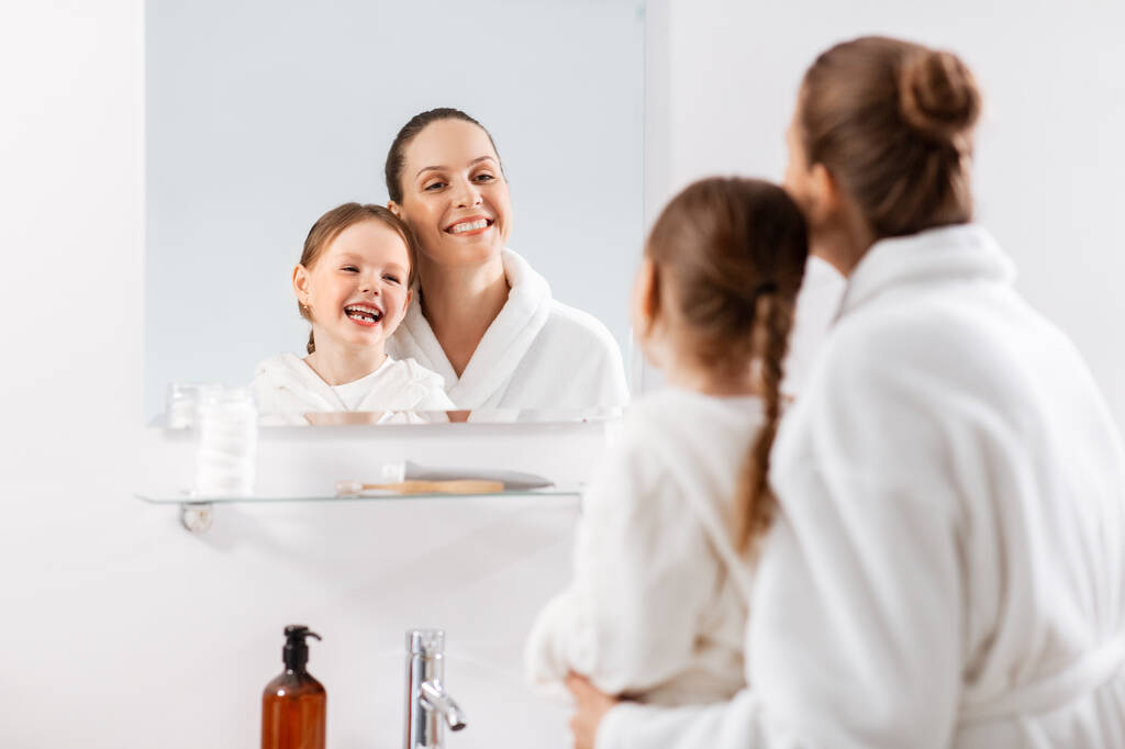 beauty, family and people concept - happy smiling mother and little daughter looking to mirror and hugging in bathroom - Photo, Image