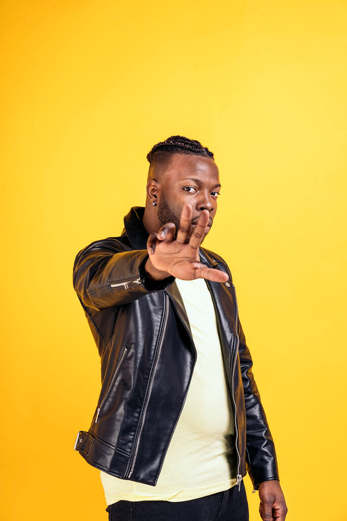 Confident black man wearing black leather jacket posing in studio shot and looking at camera against yellow background. - Zdjęcie, obraz