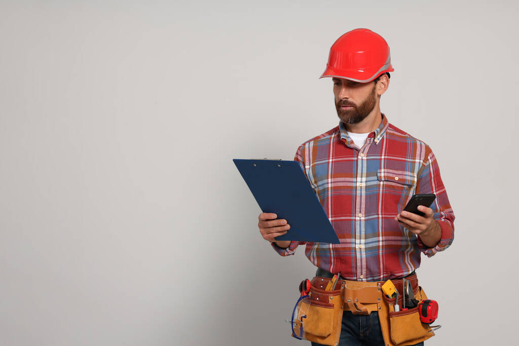 Professional builder in hard hat with clipboard, phone and tool belt on light background, space for text - Foto, immagini