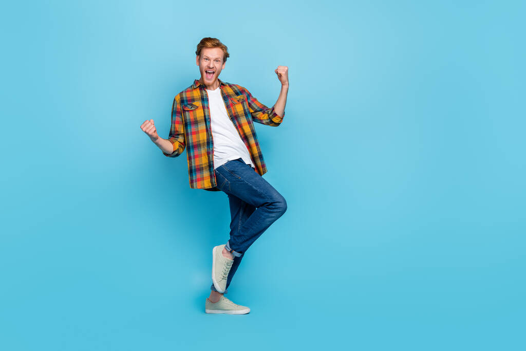 Full length photo of young satisfied man wear plaid shirt denim jeans screaming yes clenches fists isolated on blue color background. - Photo, Image