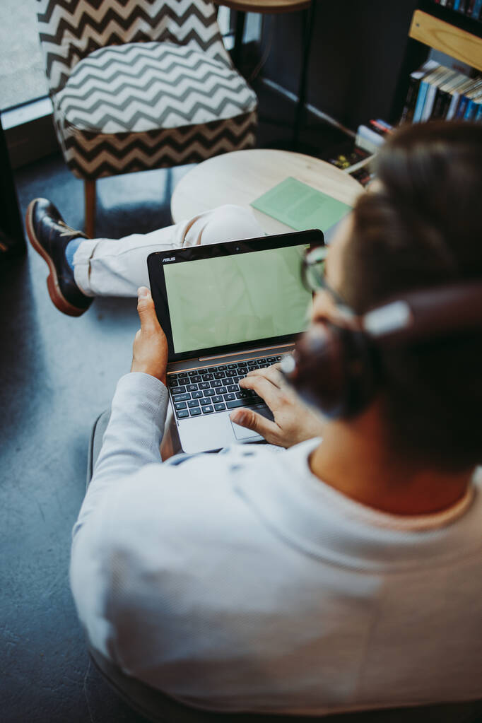 Hombre guapo de mediana edad sentado en la librería moderna, leyendo y usando su computadora portátil o portátil mientras escucha música con auriculares. - Foto, Imagen