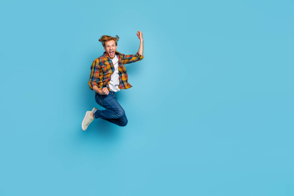 Photo of astonished cheerful guy with red hair dressed checkered shirt jumping raise fist up scream yeah isolated on blue color background. - Foto, immagini