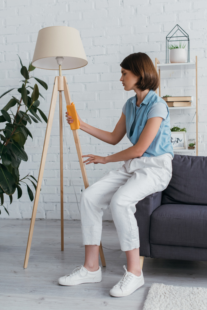 full length of woman sitting on couch and cleaning floor lamp - Foto, immagini