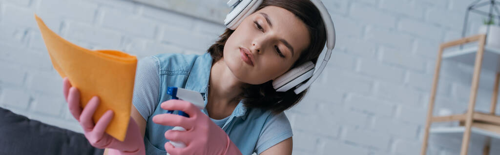 brunette woman spraying cleanser on rag while listening music in wireless headphones, banner - Foto, Bild