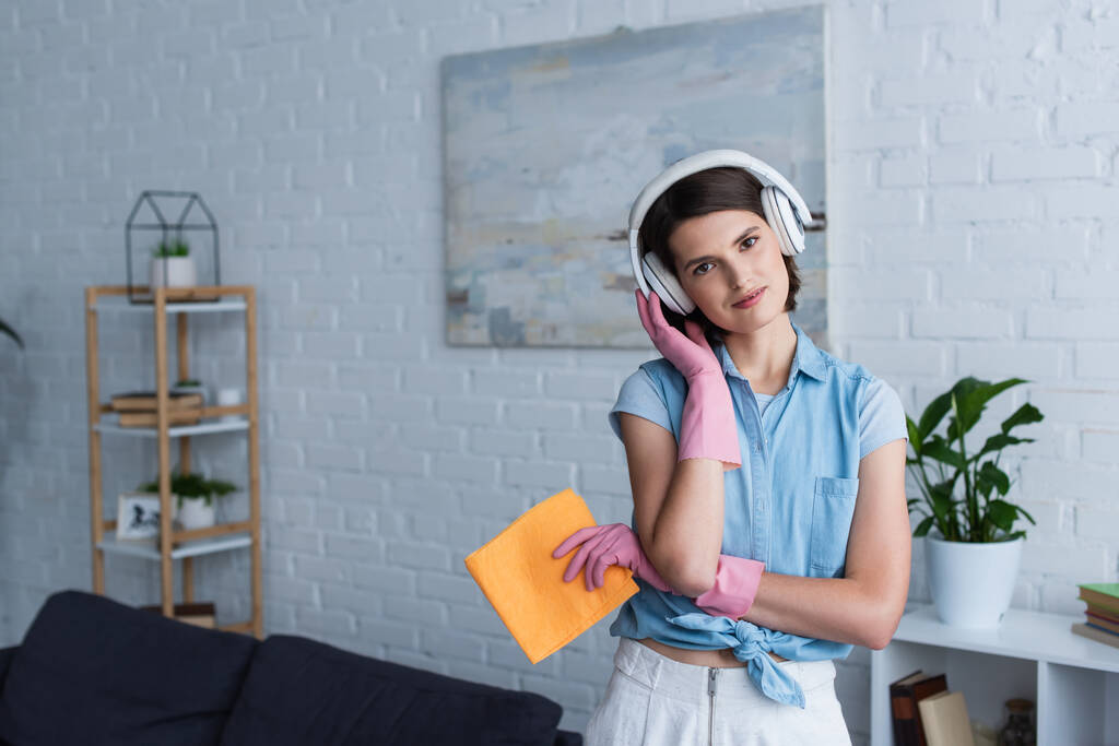 positive housewife in rubber gloves and wireless headphones holding rag and looking at camera - Fotoğraf, Görsel