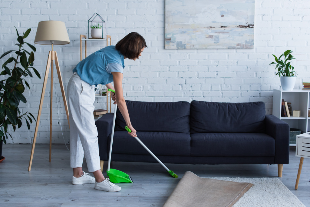 side view of woman sweeping floor near sofa in living room - Photo, Image