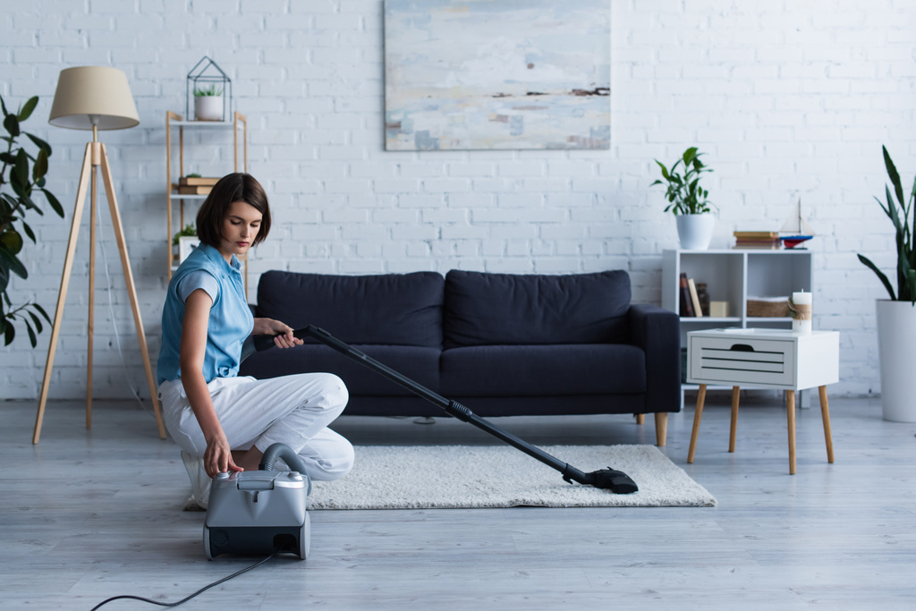Brunette woman switching vacuum cleaner near carpet in living room  - Foto, Imagem