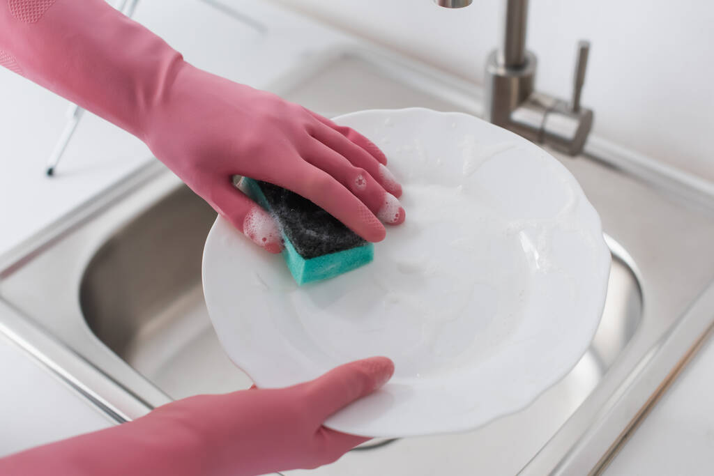 Обрезанный вид женщины в резиновых перчатках, моющая тарелка на кухне  - Фото, изображение