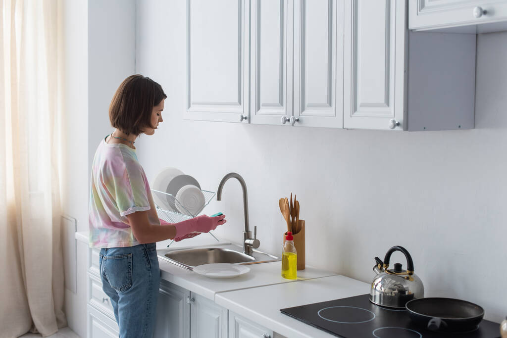 Side view of woman cleaning plate near dishwashing liquid in kitchen  - Valokuva, kuva