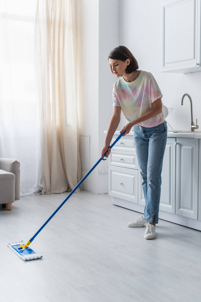 Brunette woman in casual clothes cleaning floor with mop in kitchen  - Foto, Imagen