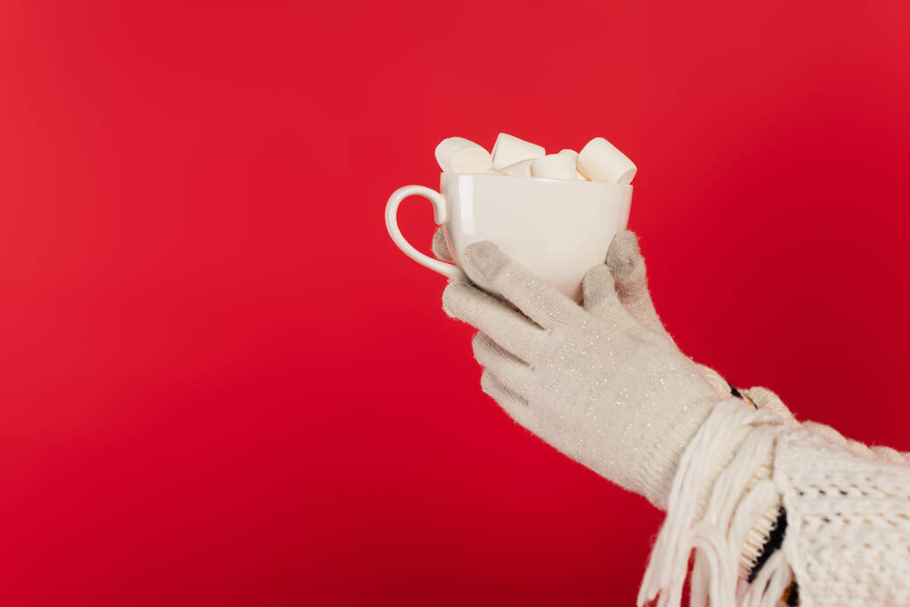 Cropped view of woman in warm scarf and gloves holding cup with marshmallows isolated on red  - Fotó, kép