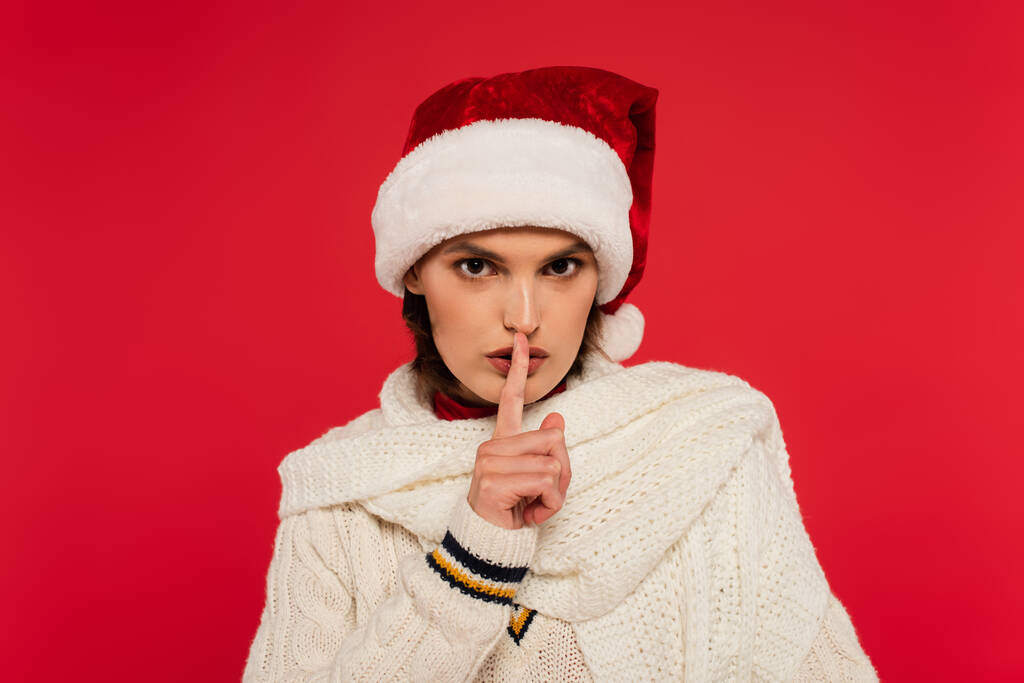 Young woman in santa hat and knitted scarf showing secret gesture isolated on red  - Foto, Imagem
