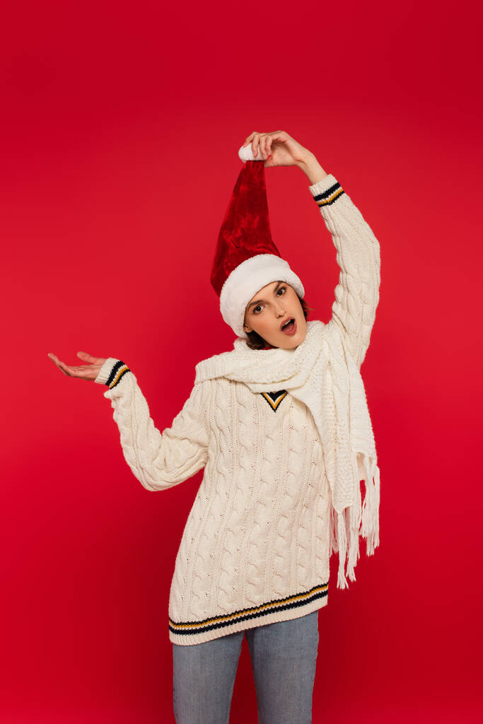 Σοκαρισμένη γυναίκα με πουλόβερ και καπέλο Σάντα δείχνει με το χέρι απομονώνονται σε κόκκινο  - Φωτογραφία, εικόνα