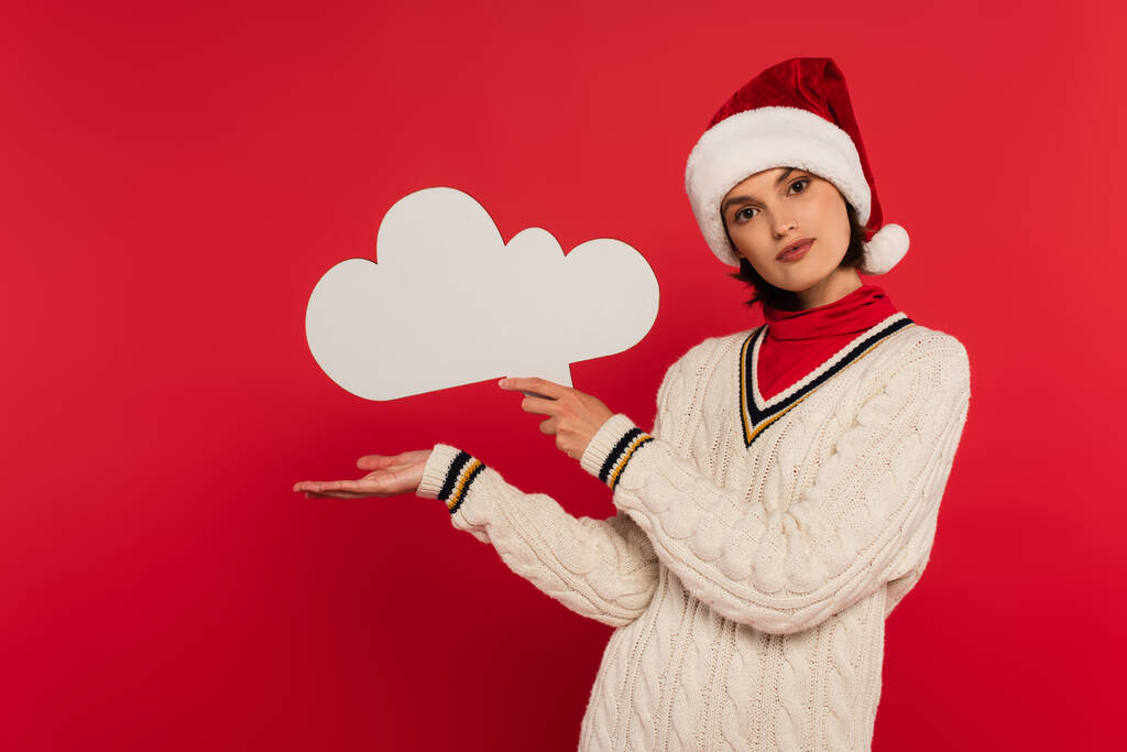 young woman in santa hat and sweater holding thought bubble and pointing with hand isolated on red - Photo, Image