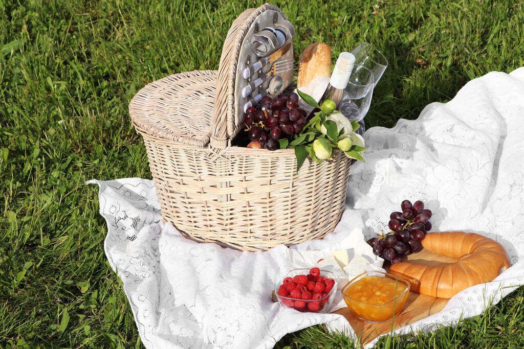 Picnic blanket with tasty food, flowers, basket and cider on green grass outdoors - Fotó, kép
