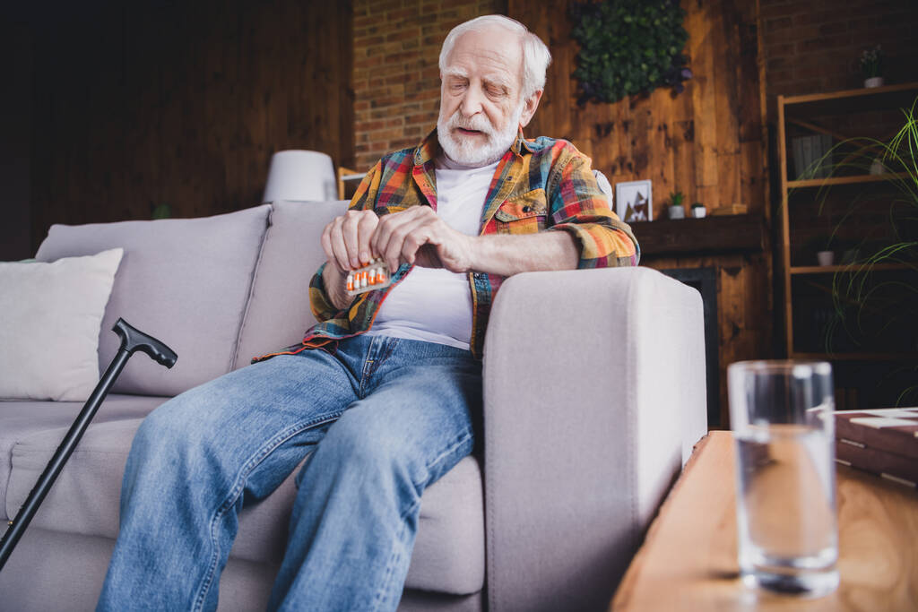 Photo of unhappy sad man pensioner wear checkered shirt drinking medicines sitting sofa indoors house room. - Fotoğraf, Görsel