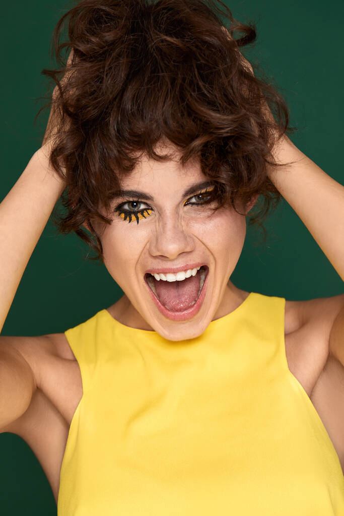 portrait of happy exited pretty young womanwith fun make-up in yellow dress starring with opened mouth on green background - Foto, Bild