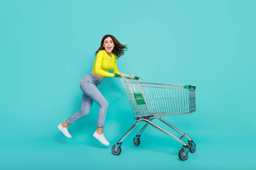 Full body photo of funny brunette hair millennial lady jump with trolley wear neon shirt jeans shoes isolated on teal color background. - Foto, immagini