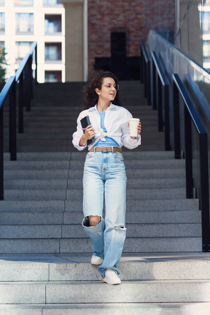 Young cheerful student woman in glasses goes down the stairs with cup of coffee and notebook in her hand. Cityspace. - Foto, imagen