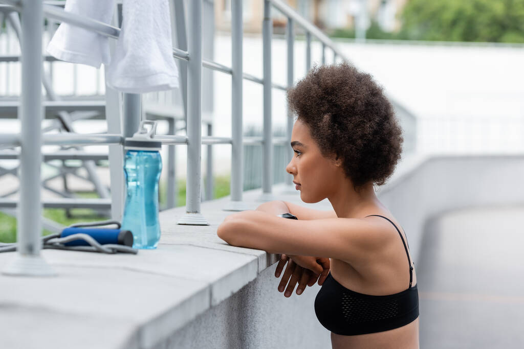 side view of brunette african american sportswoman with crossed arms near sports bottle and blurred jump rope - Foto, afbeelding