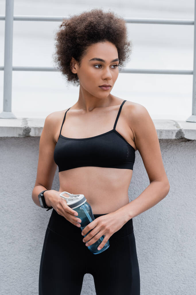 pretty african american woman in black sports bra holding sports bottle and looking away - Foto, afbeelding