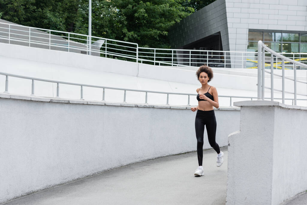полная длина стройная африканская американка в черной спортивной одежде бегает на открытом воздухе - Фото, изображение
