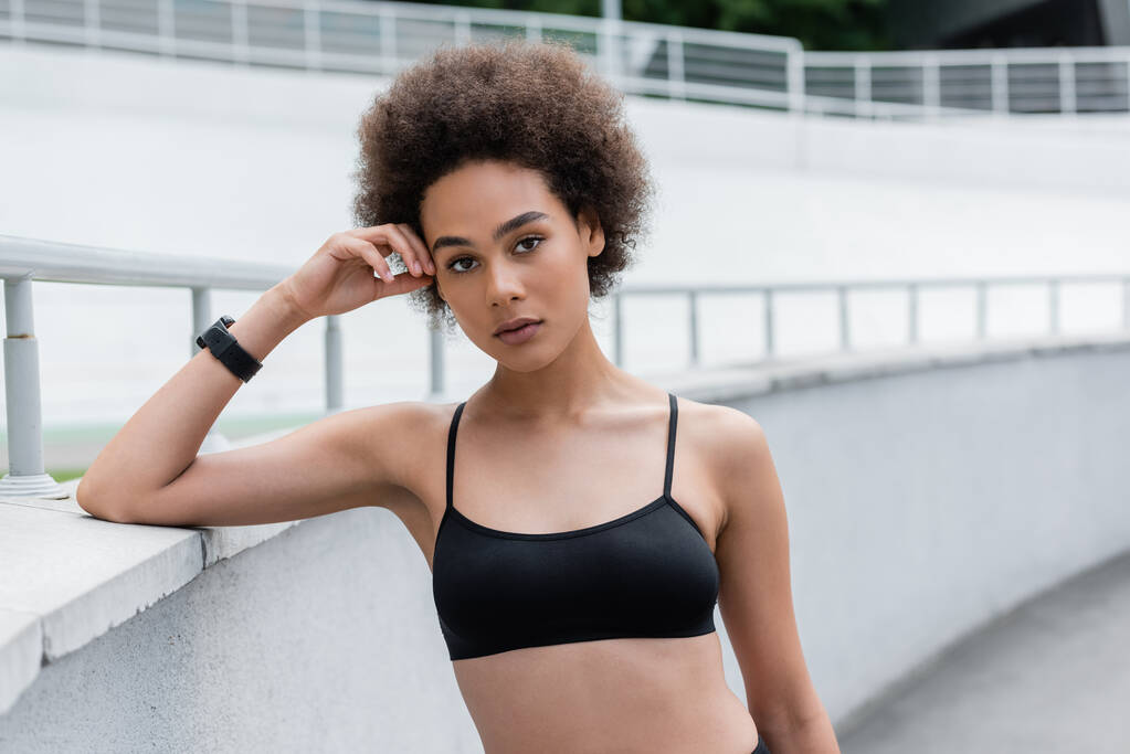 athletic african american woman in black sports bra standing with hand near head - Foto, afbeelding