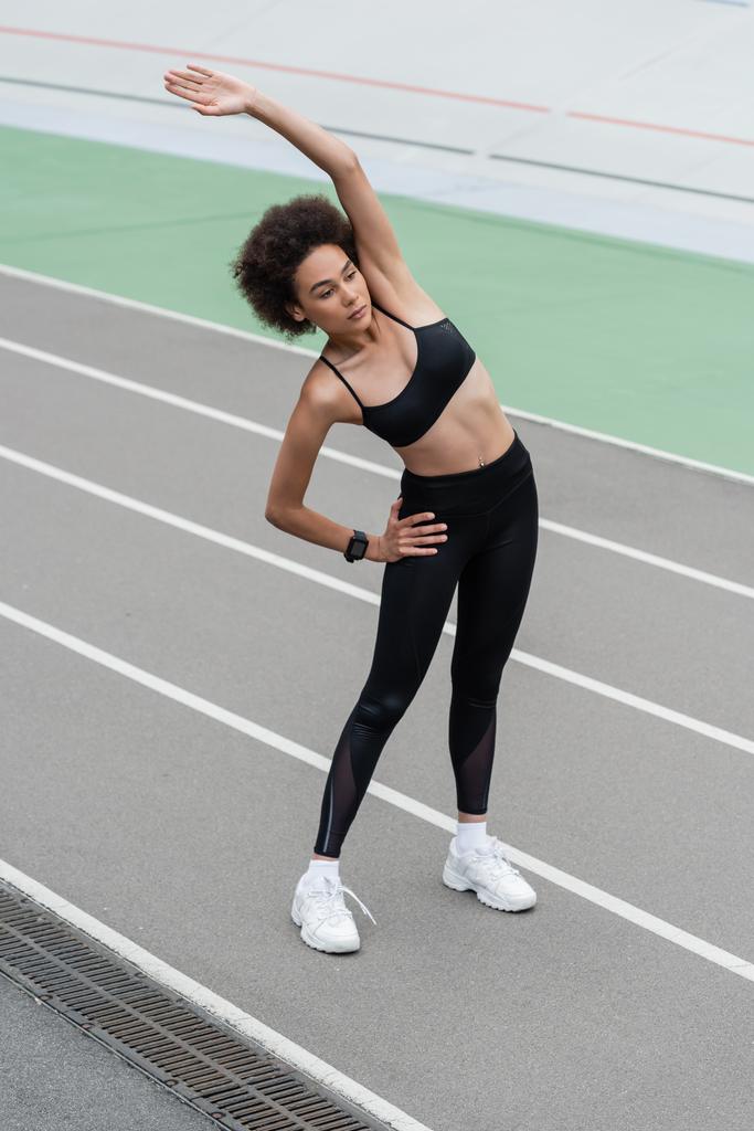 athletic african american woman in black sportswear doing side bend exercise with hand on hip and raised hand - Foto, Bild