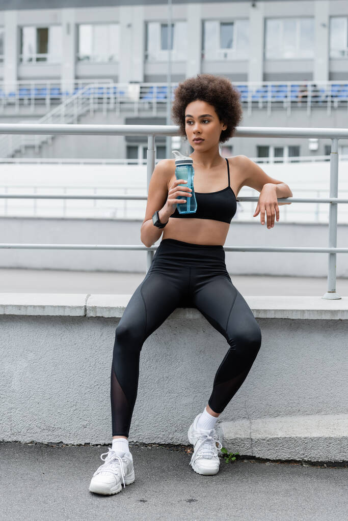 full length of african american woman in black sportswear and white sneakers sitting with sports bottle - 写真・画像