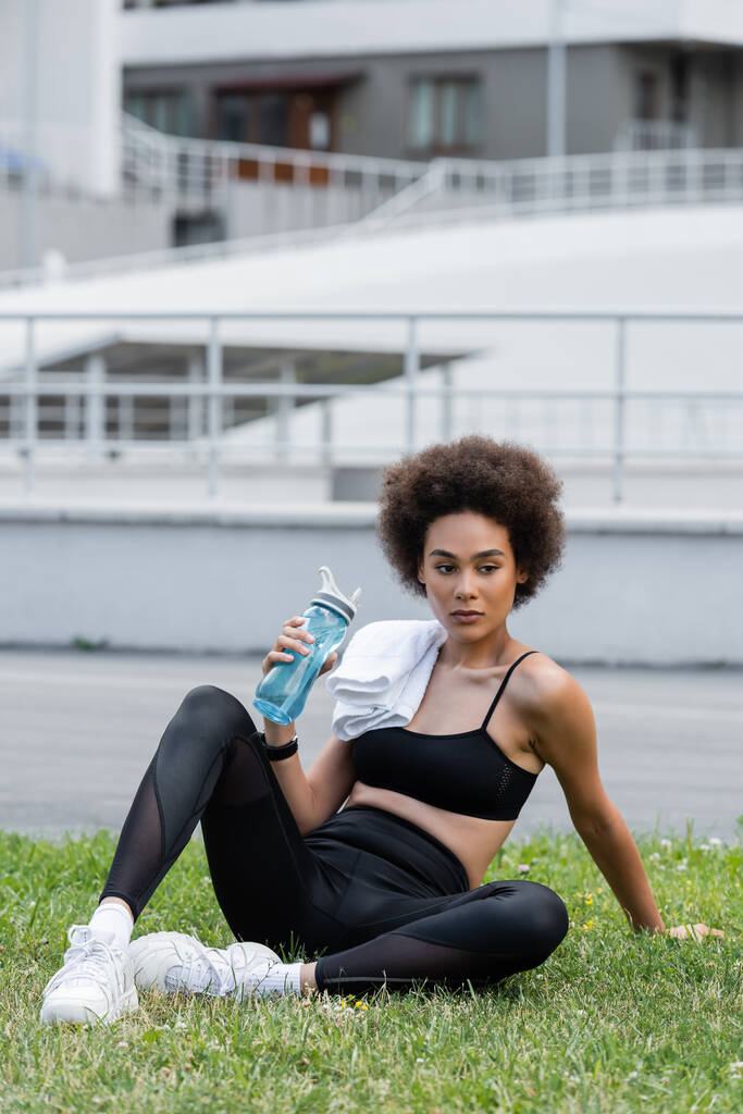slender african american woman in black sportswear sitting on lawn with towel and sports bottle - Foto, Imagem