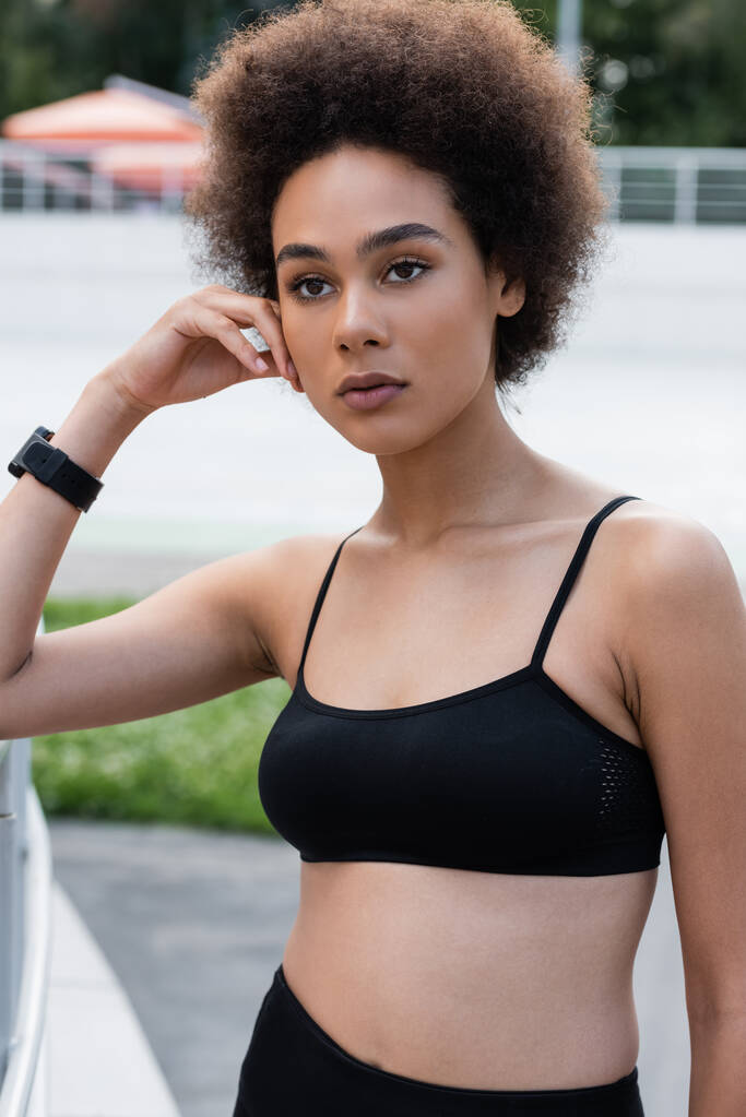 young african american woman in black sports bra and fitness tracker standing with hand near face - Foto, Bild