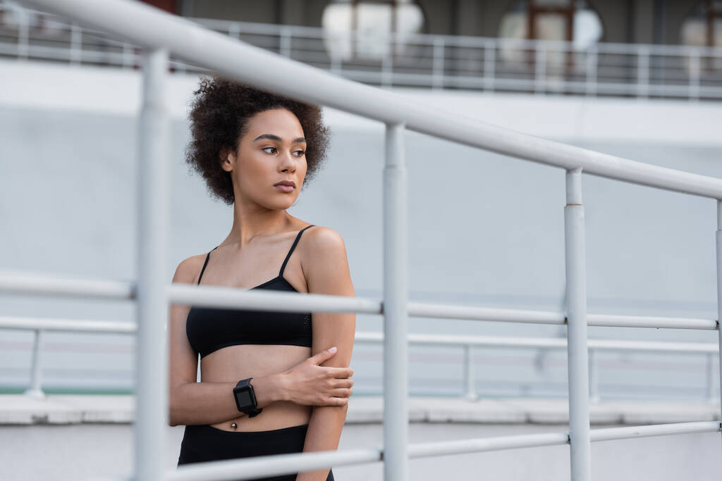 curly african american woman in black sports bra and fitness tracker standing near fence - Zdjęcie, obraz