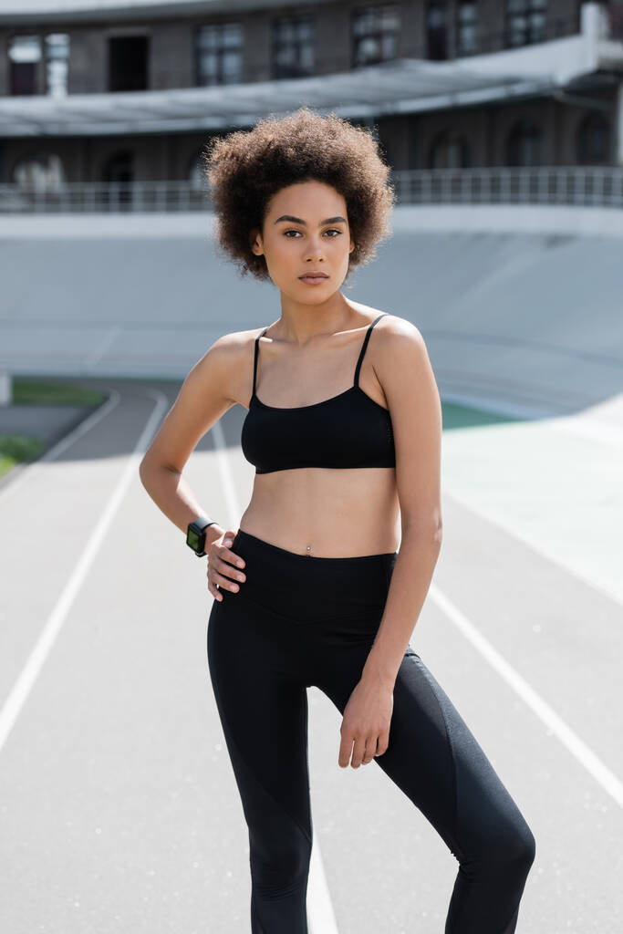 slim african american woman in black sportswear standing with hand on hip and looking at camera - Foto, afbeelding