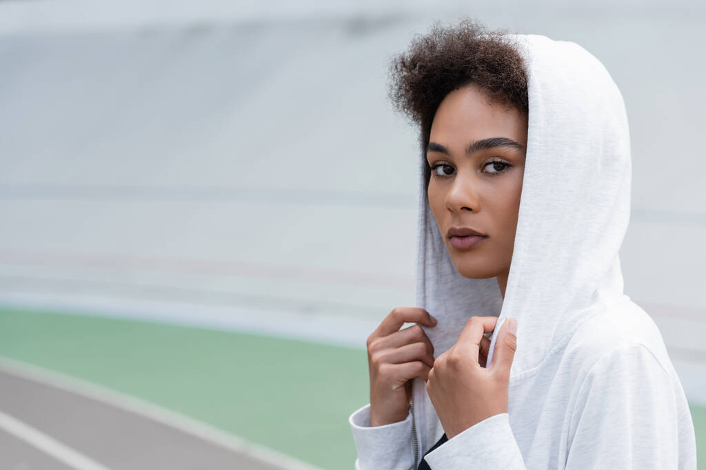 pretty african american sportswoman in white hoodie looking at camera on blurred stadium - Photo, Image