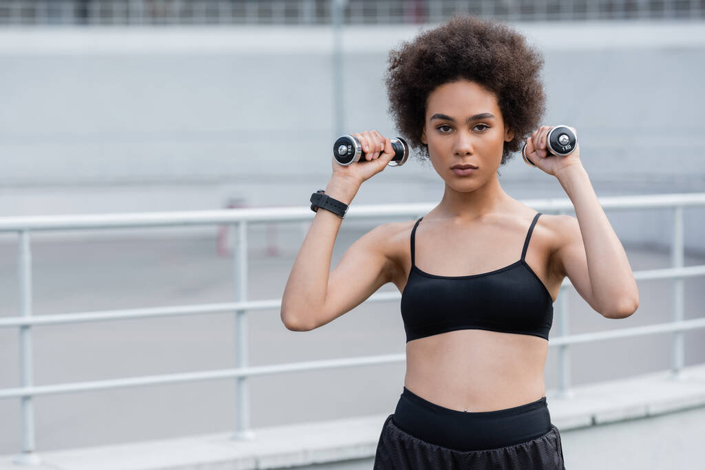 African american sportswoman training with dumbbells and looking at camera on stadium  - Foto, Imagen