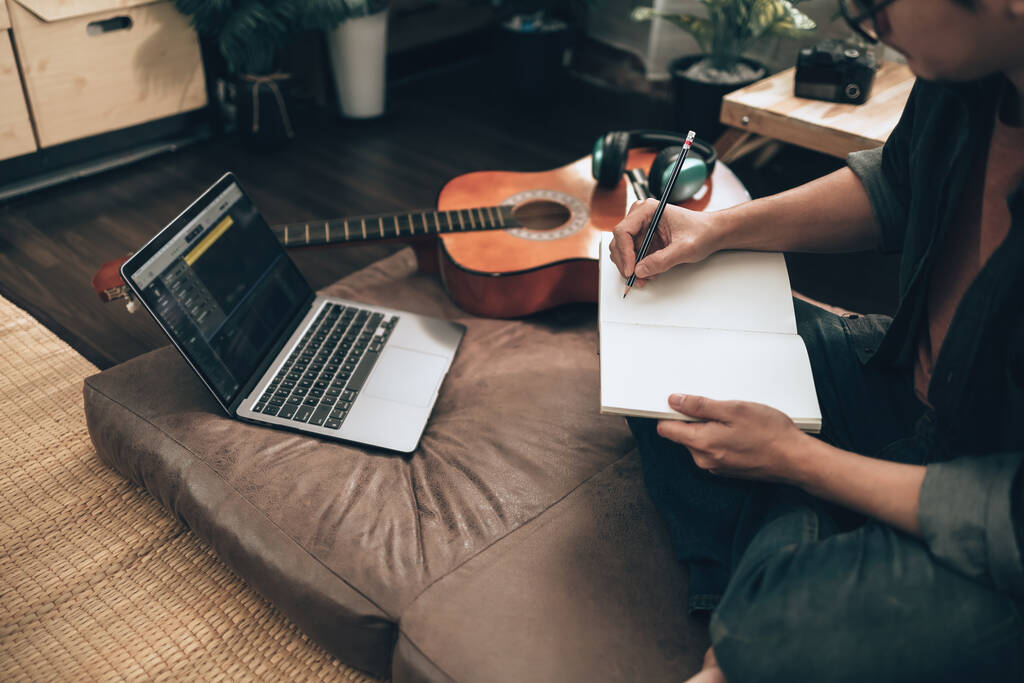 молодий чоловік розслабляється і грає на гітарі, сидячи на дивані у вітальні вдома. Музика створює мелодійну пісню, текст на ноутбуці та концепцію практики
. - Фото, зображення
