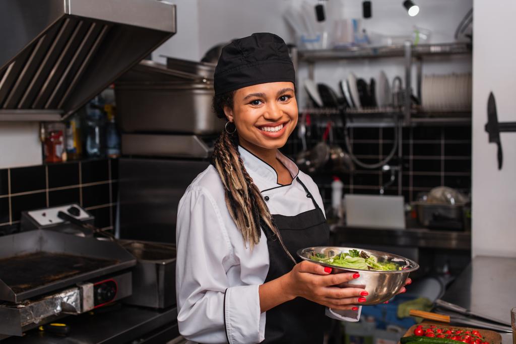 happy and young african american chef holding bowl with salad in professional kitchen - 写真・画像