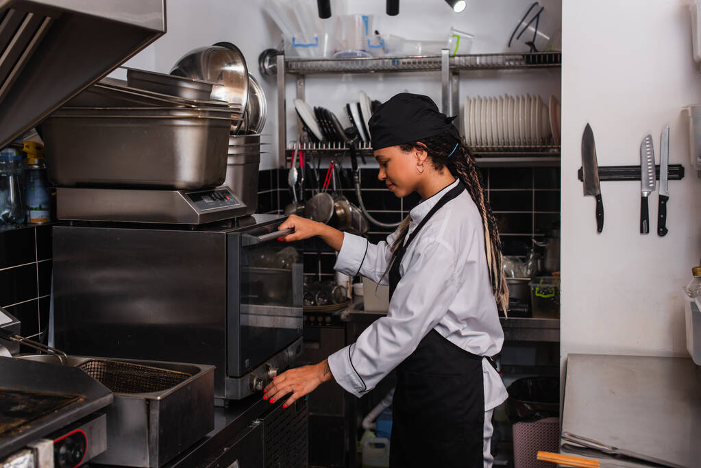 вид збоку молодого афроамериканського шеф-кухаря в капелюсі, що стоїть біля конвекційної печі на кухні
  - Фото, зображення